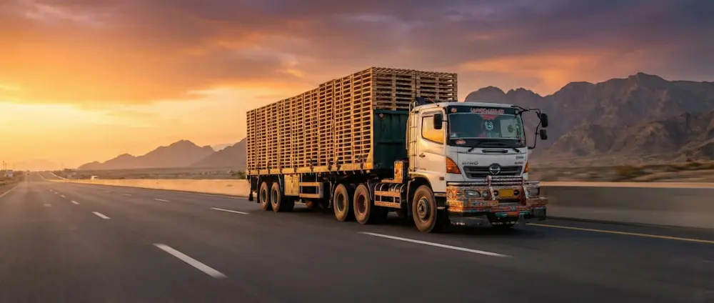 A large flatbed semi-truck fully loaded with brand new wooden pallets driving at speed on a highway during sunset, illustrating nationwide logistics and reliable delivery.